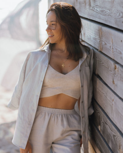 A woman wearing a beige linen crossover cropped top with wide straps and a cross over front detail. She is also wearing light-colored pants and is standing next to a wooden structure.