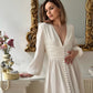 A woman in a white plunged long sleeve maxi dress, standing in a room with a gold-frame mirror and a floral arrangement in the background.