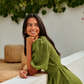 A woman  wearing a sage green linen dress leans against a white wall in a sunlit outdoor setting with green foliage overhead.