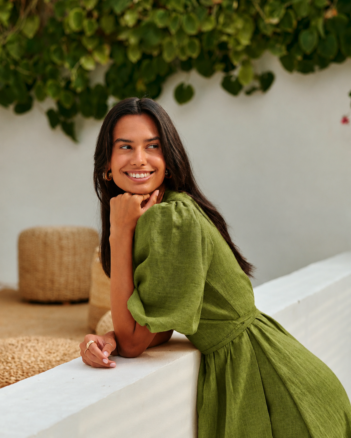 A woman  wearing a sage green linen dress leans against a white wall in a sunlit outdoor setting with green foliage overhead.