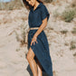 Woman in a navy dress standing on sandy terrain with sparse vegetation.