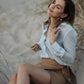A woman sitting on a sandy beach wearing a sky blue silk shirt with a classic collar and long sleeves, buttoned cuffs, with the shirt's front button fastening visible.