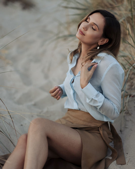 A woman sitting on a sandy beach wearing a sky blue silk shirt with a classic collar and long sleeves, buttoned cuffs, with the shirt's front button fastening visible.