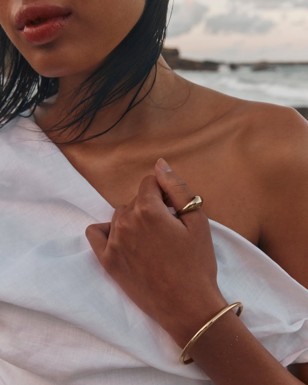 Close-up detail of a white one shoulder linen dress with soft draping, gold jewelry, and lightweight textured linen fabric.