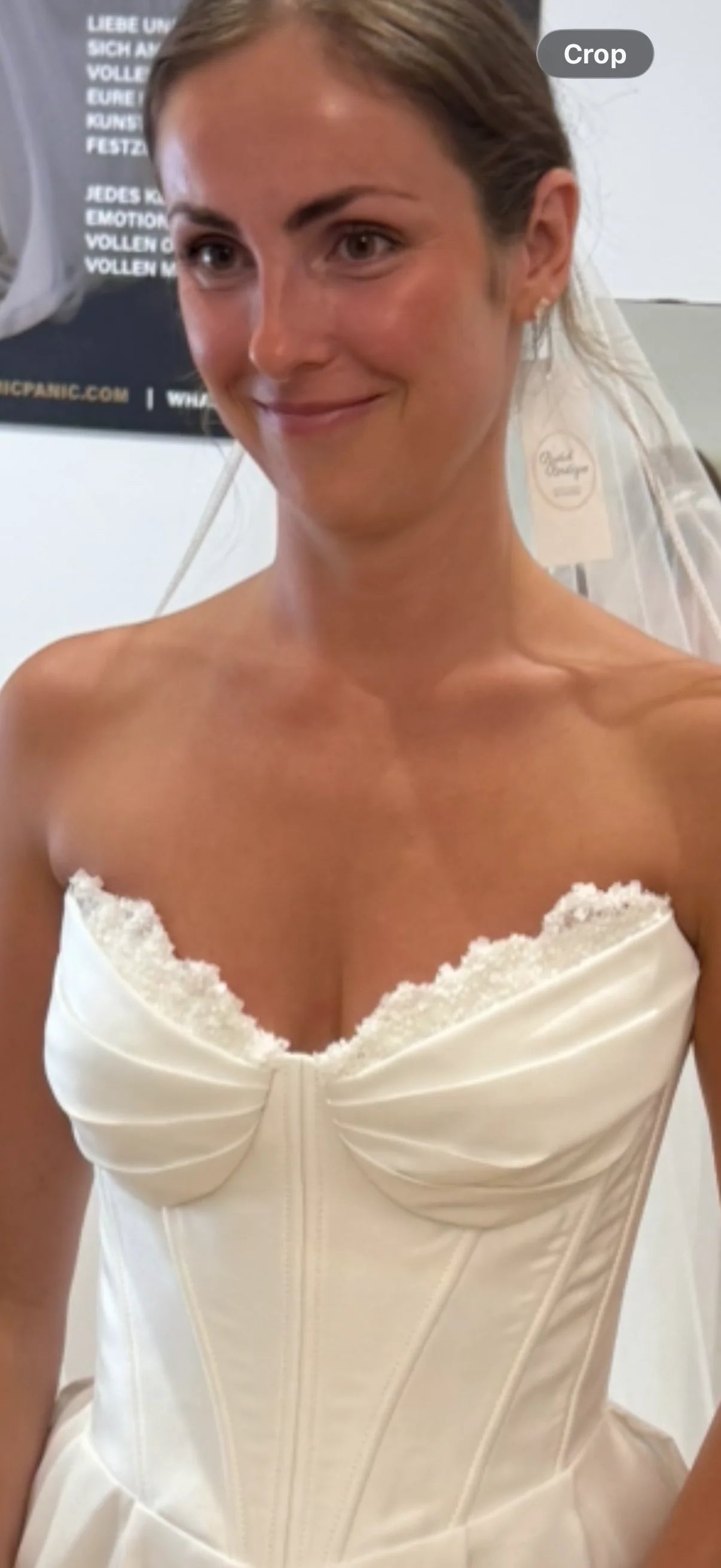 A woman wearing a strapless white wedding dress with a sweetheart neckline, lace trim, and corset-style bodice, photographed indoors against a light background.