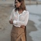 Woman standing on a beach wearing a white blouse and brown skirt.