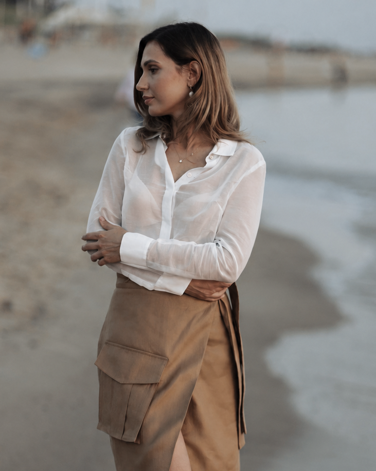 Woman standing on a beach wearing a white blouse and brown skirt.