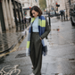 A woman stands on a wet city street wearing a blue, gray, and lime green striped knit scarf, a light blue top, olive green wide-leg trousers, and beige shoes.