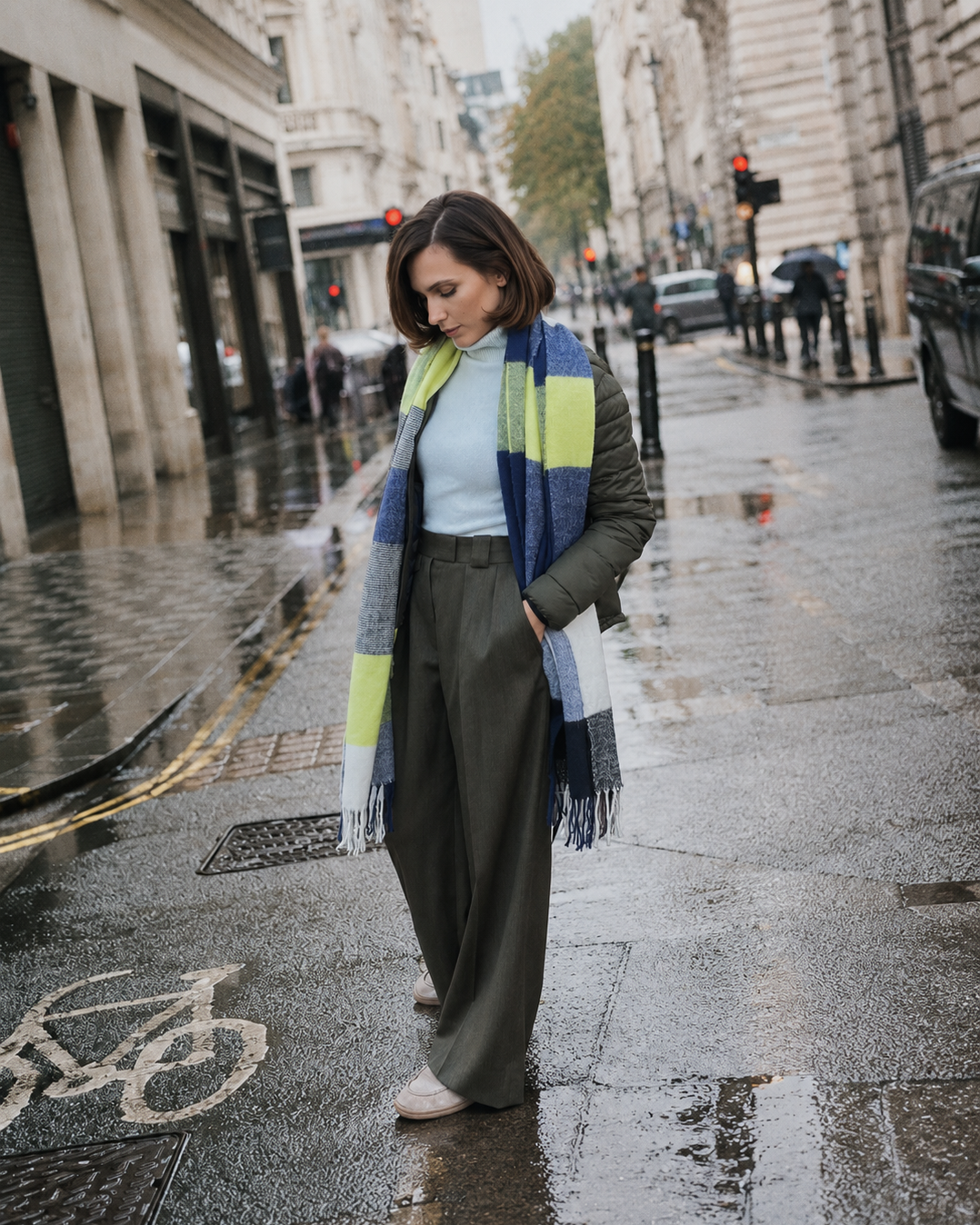 A woman stands on a wet city street wearing a blue, gray, and lime green striped knit scarf, a light blue top, olive green wide-leg trousers, and beige shoes.