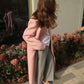 A woman stands outdoors in front of a building wall and flowerbeds, wearing a long pink coat over a white top and gray straight-leg pants with white pointed flats.