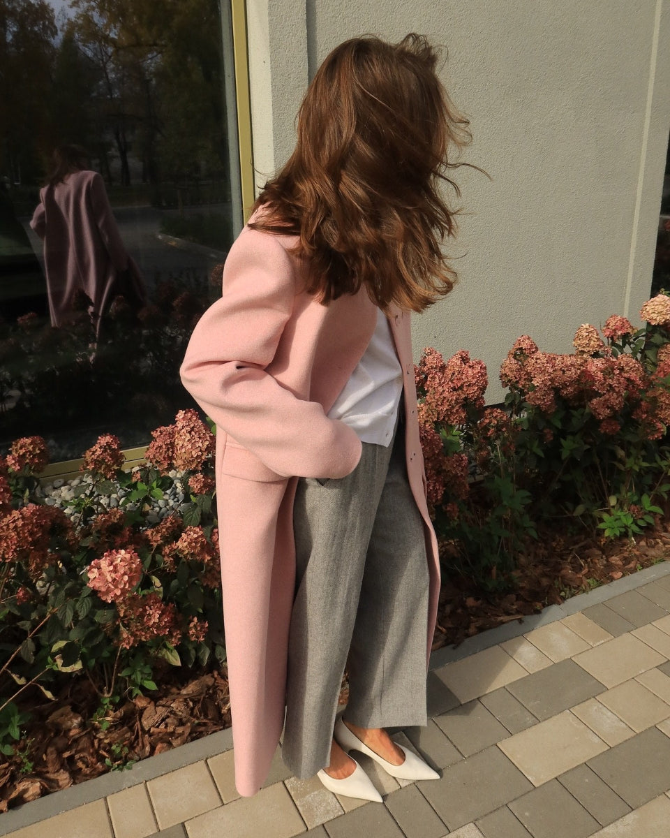 A woman stands outdoors in front of a building wall and flowerbeds, wearing a long pink coat over a white top and gray straight-leg pants with white pointed flats.