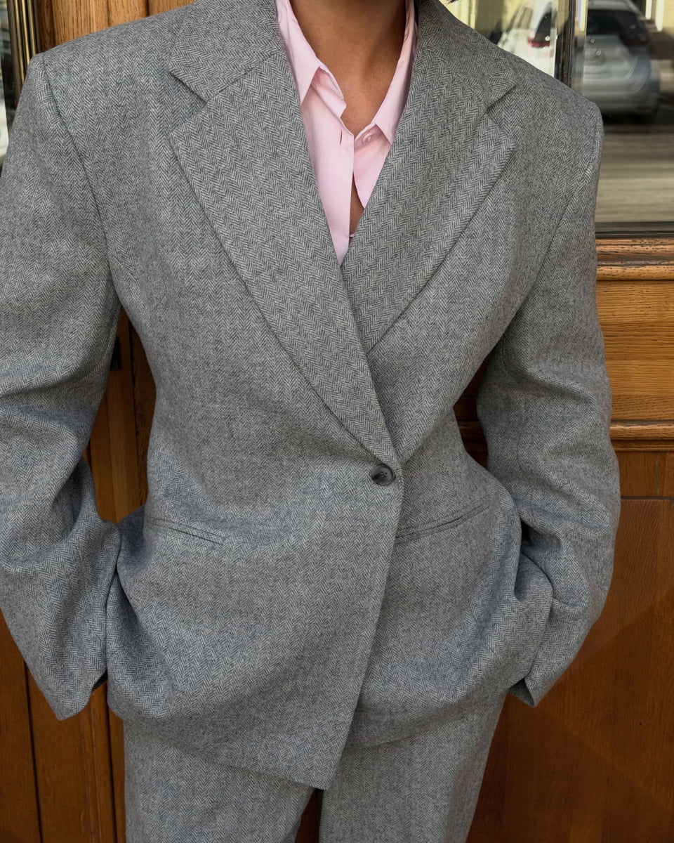 A person wearing a tailored wool gray blazer and matching trousers over a light pink collared shirt, standing in front of a wooden door.