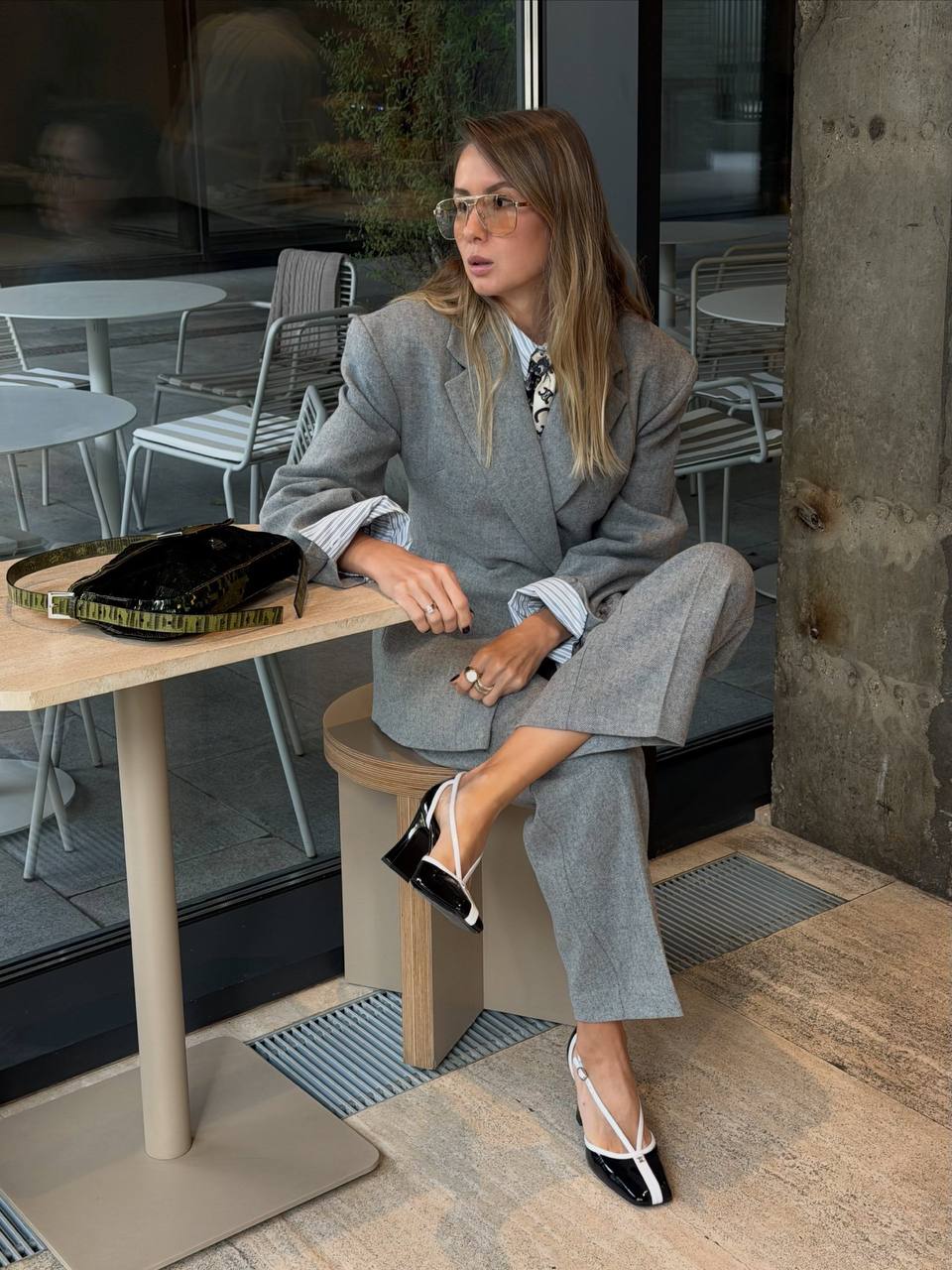 A woman sits on a stool in a cafe setting wearing a matching light gray tailored wool suit with wide lapels and straight legs, layered over a white collared shirt. She is wearing black and white strappy heels and holding a black patterned clutch.