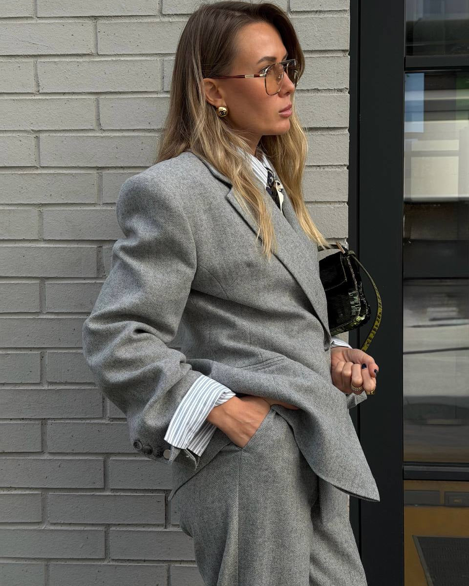 A woman wearing a tailored light gray suit with a matching blazer and trousers, styled over a white striped button-down shirt. She is standing against a gray brick wall next to a glass door, holding a black shoulder bag.