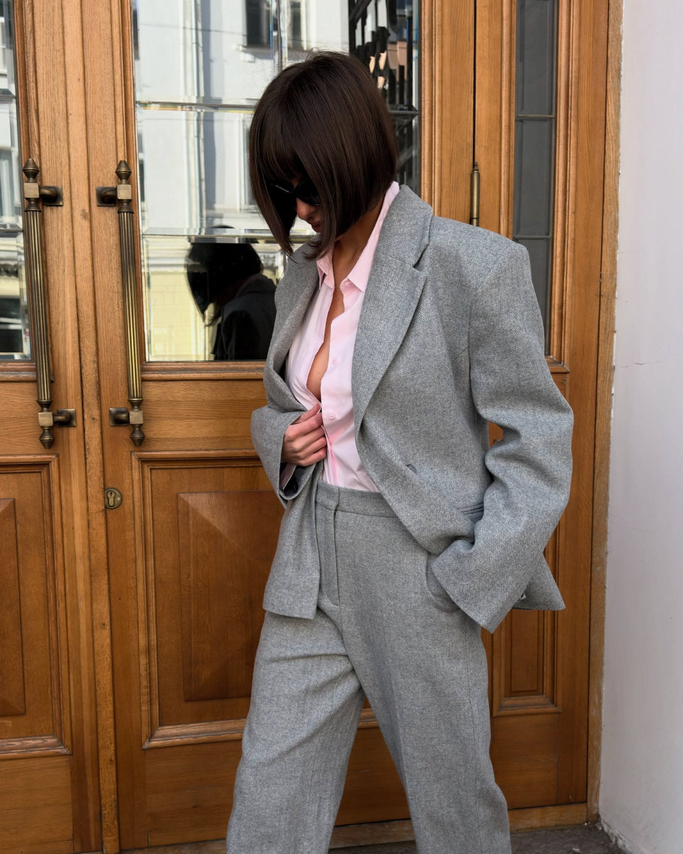 A person wearing a light gray tailored suit with a pink shirt, standing in front of a large wooden door with glass panels.