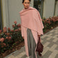 A woman stands outdoors on a paved walkway wearing a long, draped dusty pink wrap over gray straight-leg pants, with white sneakers.