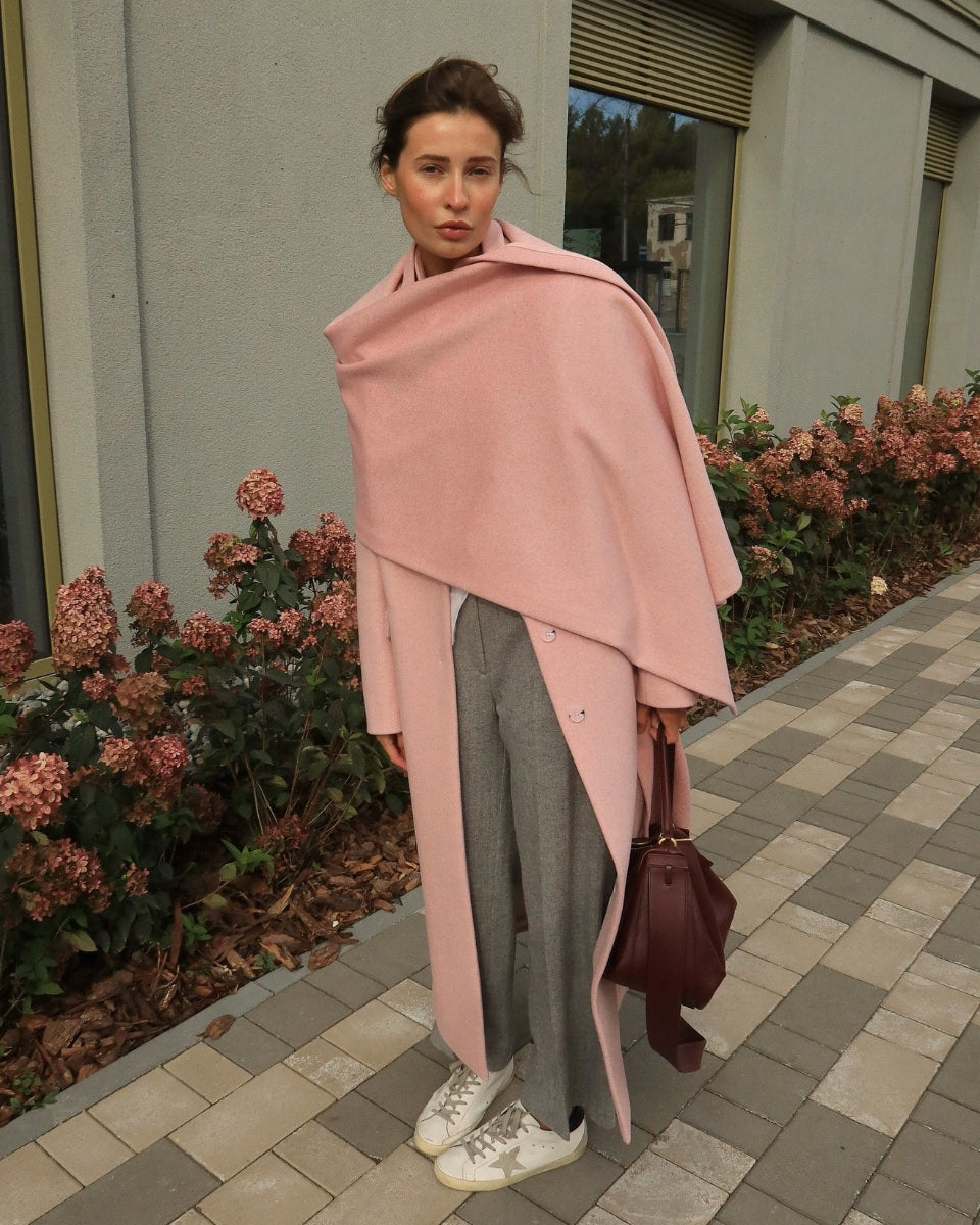 A woman stands outdoors on a paved walkway wearing a long, draped dusty pink wrap over gray straight-leg pants, with white sneakers.