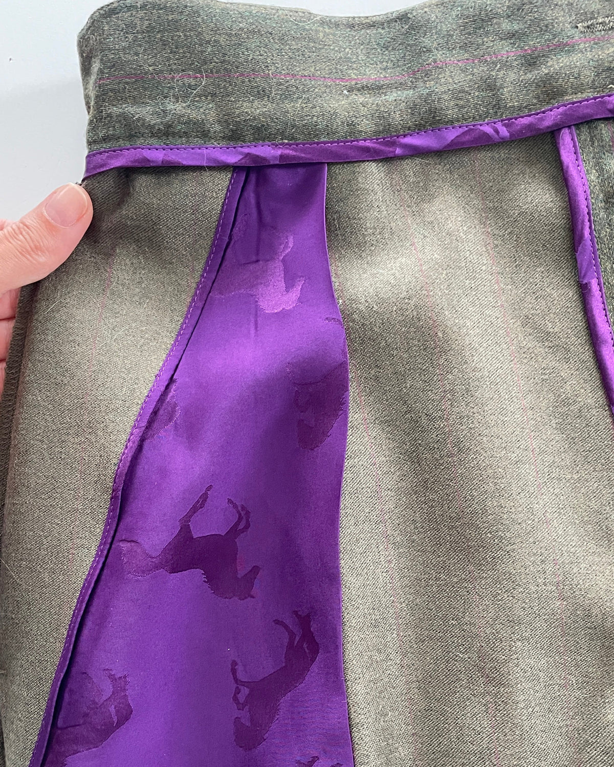 A close-up of a green textured garment with a prominent purple silk panel featuring a repeating dark horse pattern, held by a hand on the left.