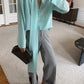 A person standing indoors wearing a long-sleeve Tiffany color slk blouse with a draped tie detail at the neckline, paired with gray straight-leg trousers and brown loafers.