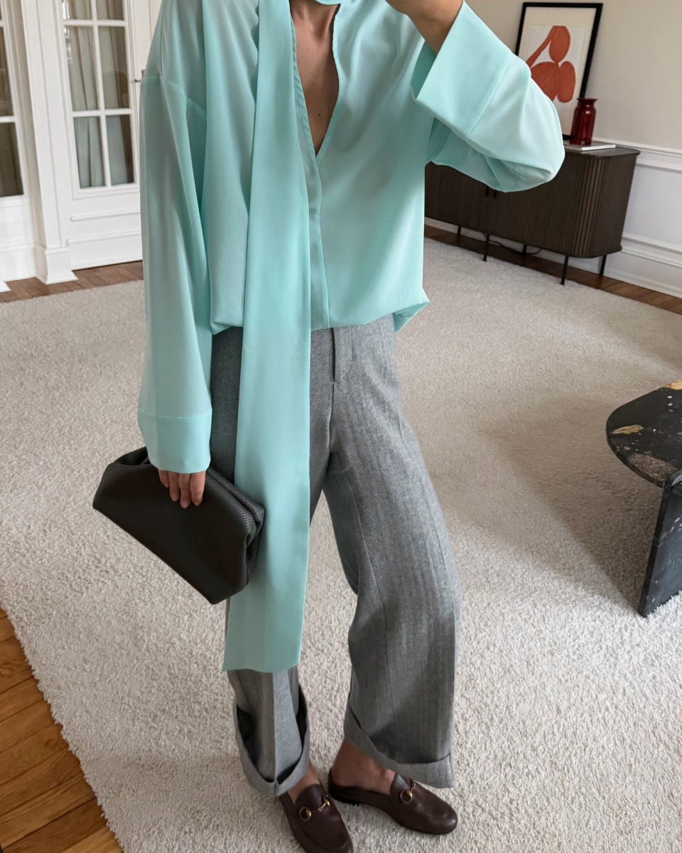 A person standing indoors wearing a long-sleeve Tiffany color slk blouse with a draped tie detail at the neckline, paired with gray straight-leg trousers and brown loafers.