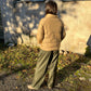 A person standing outdoors in front of a weathered wall, viewed from behind, wearing a tan quilted puffer jacket and olive green wide-leg pants.