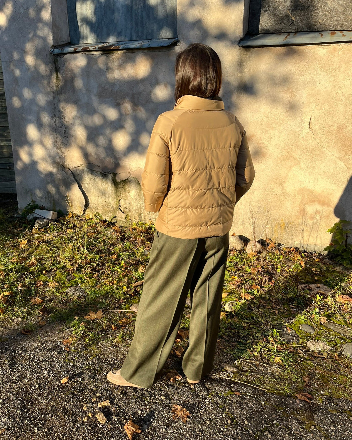 A person standing outdoors in front of a weathered wall, viewed from behind, wearing a tan quilted puffer jacket and olive green wide-leg pants.