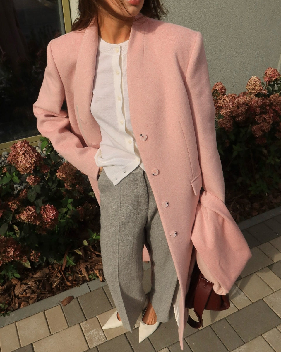 A woman wearing a light pink structured coat over a white top and gray straight-leg trousers, standing outdoors on a paved walkway with plants in the background.
