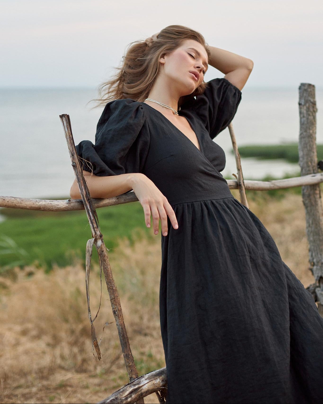 A woman wearing a black puff-sleeve midi dress leans against a rustic wooden fence outdoors near the ocean.