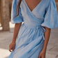 A woman wearing a sky blue linen wrap dress with puff sleeves and a tie waist, photographed outdoors in a sunlit stone setting.
