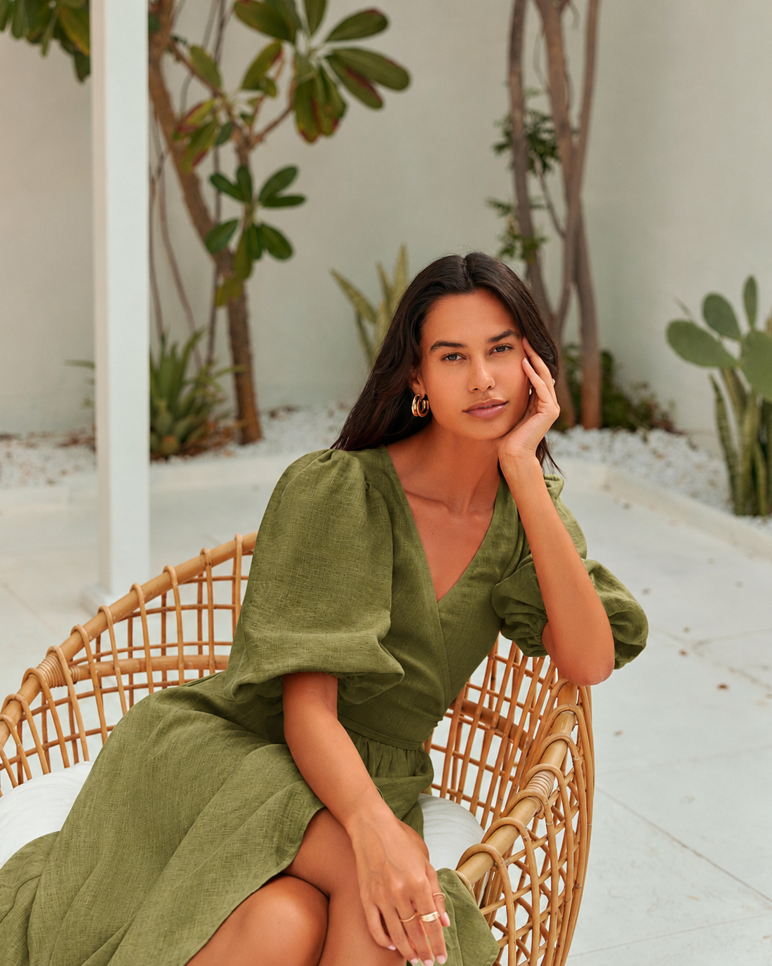 A woman wearing an moss green linen dress sits in a round woven rattan chair.