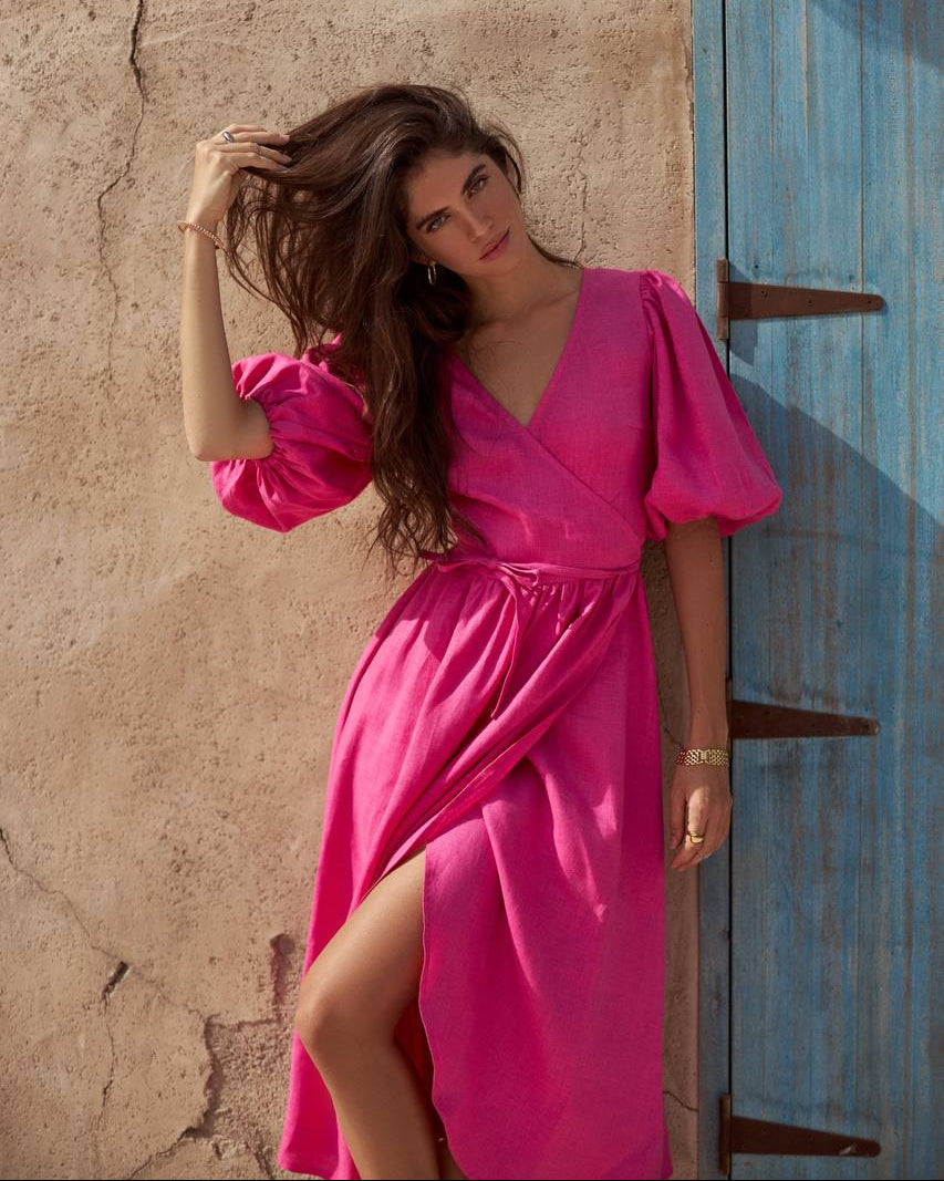 A woman wearing afuchia wrap-style midi dress with puff sleeves and a thigh-high slit.
