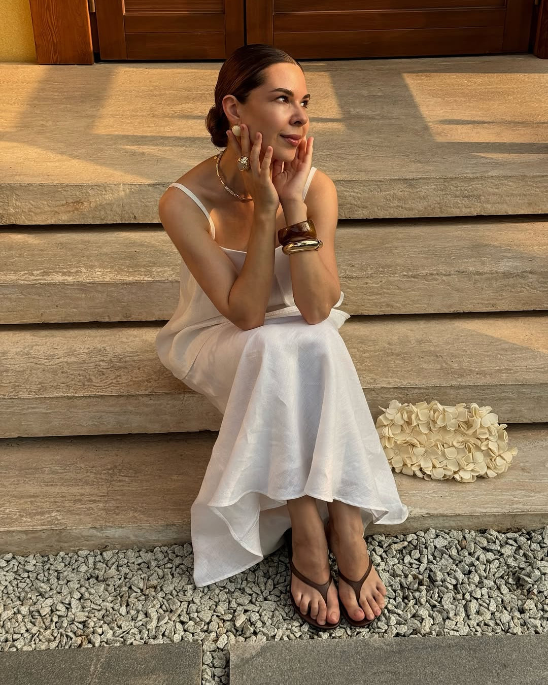 A woman wearing a white linen top and skirt  and brown sandals sits on stone steps outdoors.