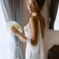 Woman standing by a window wearing an ivory backless silk maxi dress and holding calla lilies.