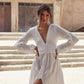 A woman wearing a white long-sleeve maxi dress with a deep V-neckline, buttoned waist, and side slit, posing outdoors in front of rustic beige stone steps and buildings.