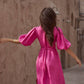 A woman viewed from behind wearing a bright pink midi dress with voluminous puff sleeves and a tie waist, standing in a narrow outdoor alley with beige stucco walls.