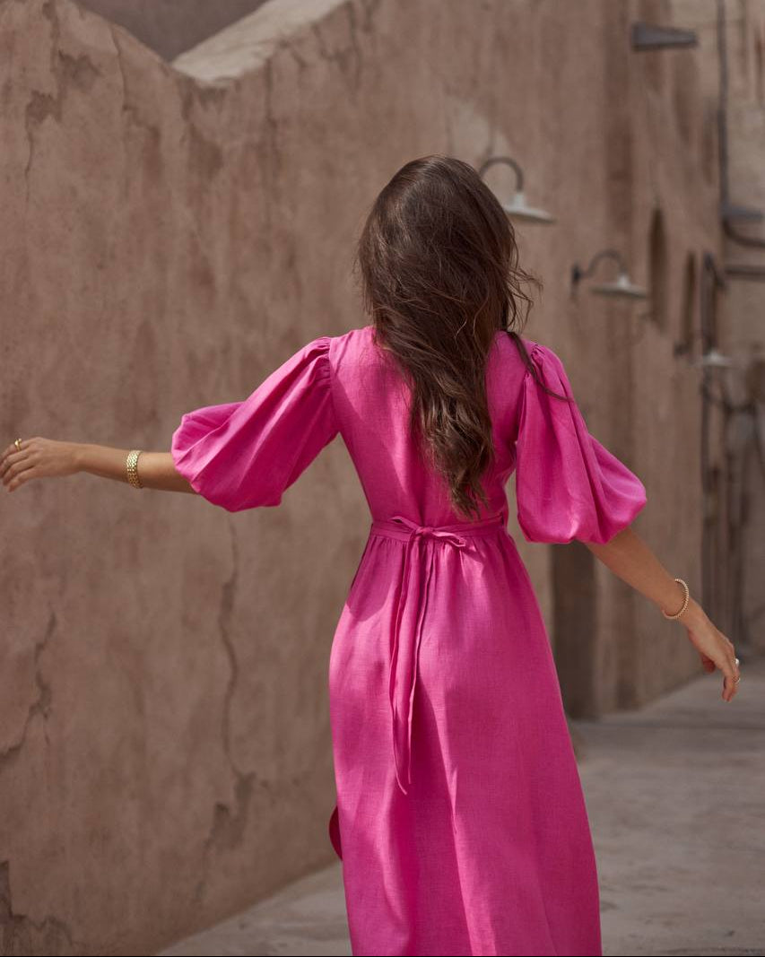 A woman viewed from behind wearing a bright pink midi dress with voluminous puff sleeves and a tie waist, standing in a narrow outdoor alley with beige stucco walls.