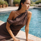 Woman sitting by a pool wearing a chocolate brown one shoulder linen dress with a draped neckline.