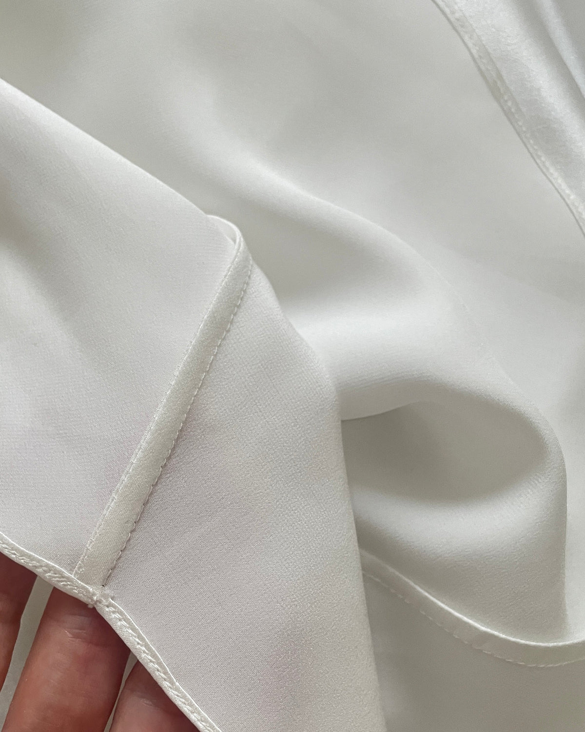 A close-up of a white silk dress inside with a hand holding the edge to show the smooth texture and fine stitching.