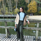 A woman stands on a wooden boardwalk by a lake wearing a beige and black puffer jacket over a light blue top and dark green wide-leg trousers, holding a small black handbag.