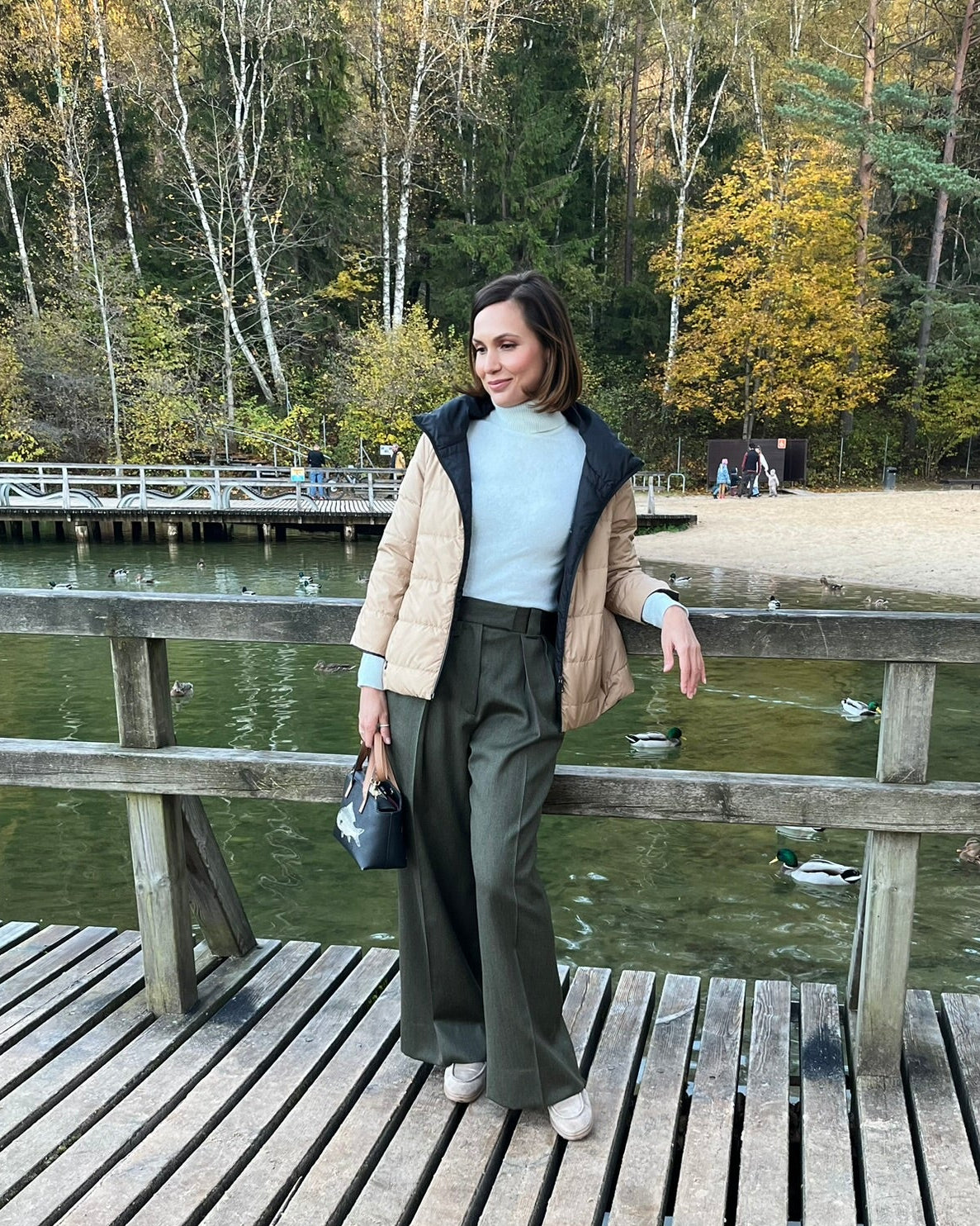 A woman stands on a wooden boardwalk by a lake wearing a beige and black puffer jacket over a light blue top and dark green wide-leg trousers, holding a small black handbag.