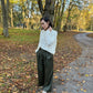 A woman stands on a leaf-strewn path in a park, wearing a white textured knit sweater and dark olive green wide-leg trousers.