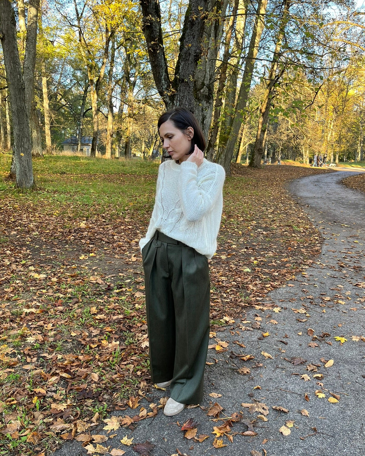 A woman stands on a leaf-strewn path in a park, wearing a white textured knit sweater and dark olive green wide-leg trousers.