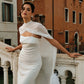 Woman in a white wedding dress wearing a sheer silk chiffon bridal cape with draping across the front, standing on a stone bridge with historic buildings behind her.