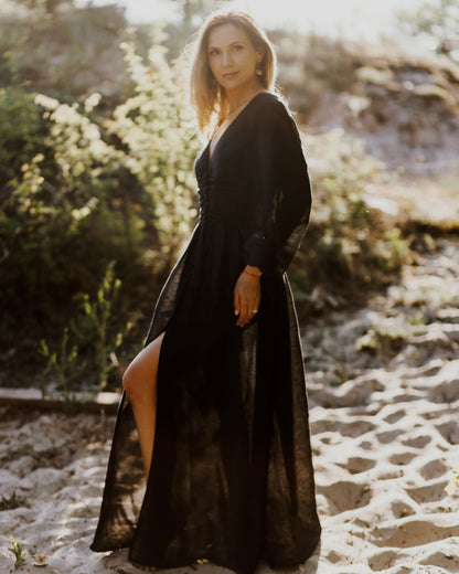 A woman stands in an elegant black dress with long sleeves and a high slit in the front against a sunset background.