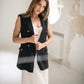 A woman wearing a black double-breasted sleeveless blazer with gold buttons over a ivory dress, standing in a bright, modern room with a textured white wall.