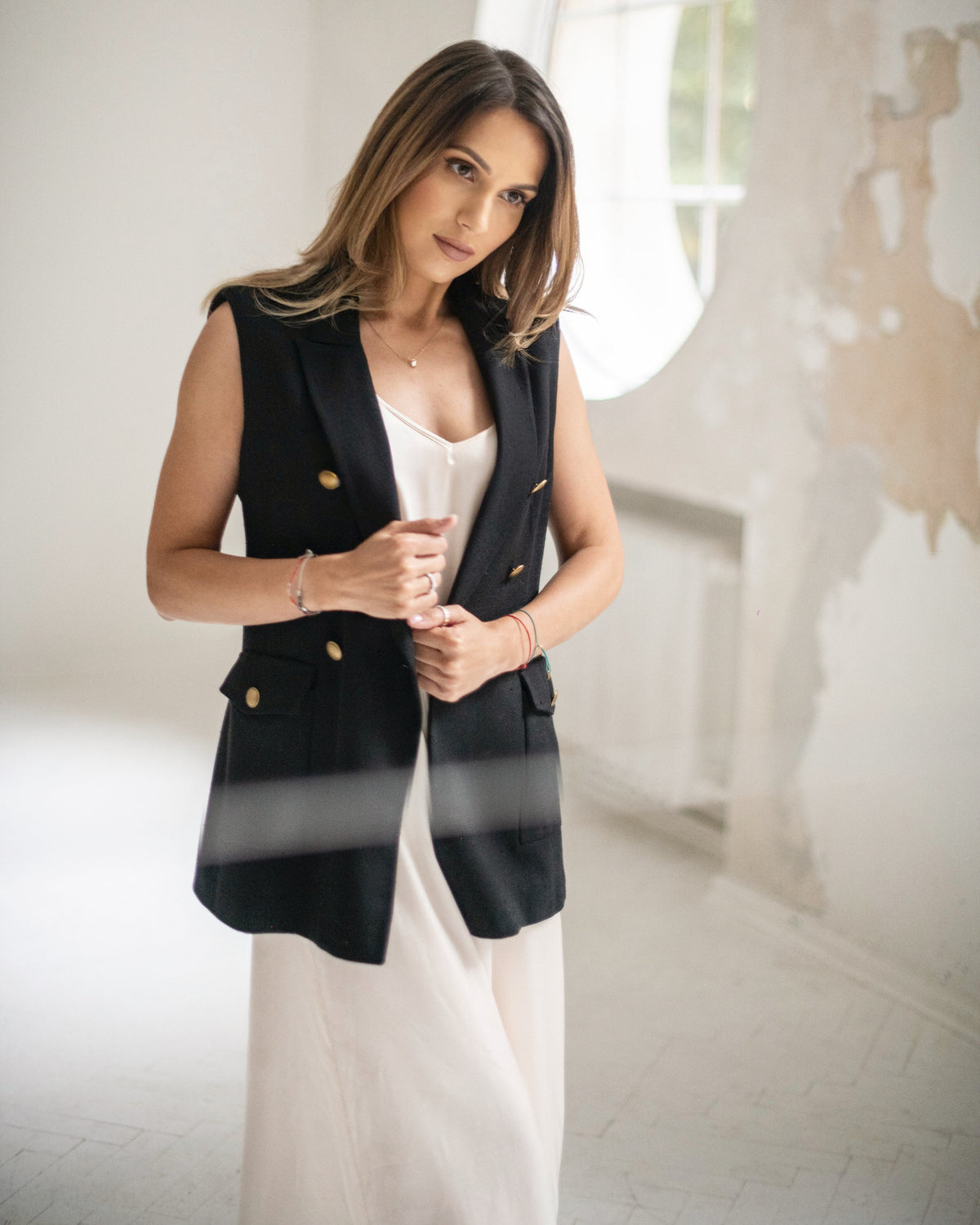 A woman wearing a black double-breasted sleeveless blazer with gold buttons over a ivory dress, standing in a bright, modern room with a textured white wall.