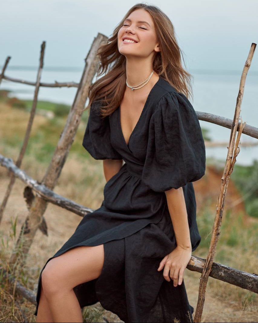 Woman in a black wrap dress sitting on a wooden fence with a scenic background