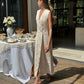 Woman wearing an natural sleeveless linen wrap maxi dress with a V-neck and defined waist, standing beside a table set with flowers and food on a sunny terrace.