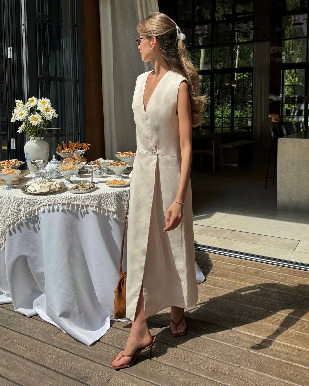 Woman wearing an natural sleeveless linen wrap maxi dress with a V-neck and defined waist, standing beside a table set with flowers and food on a sunny terrace.