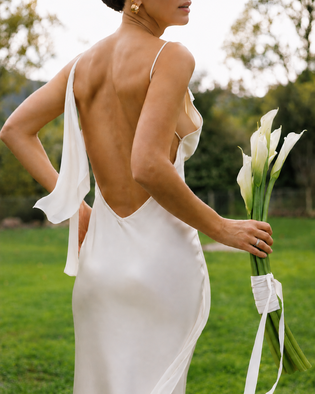 Woman holding white calla lilies while wearing an ivory open back silk maxi dress.